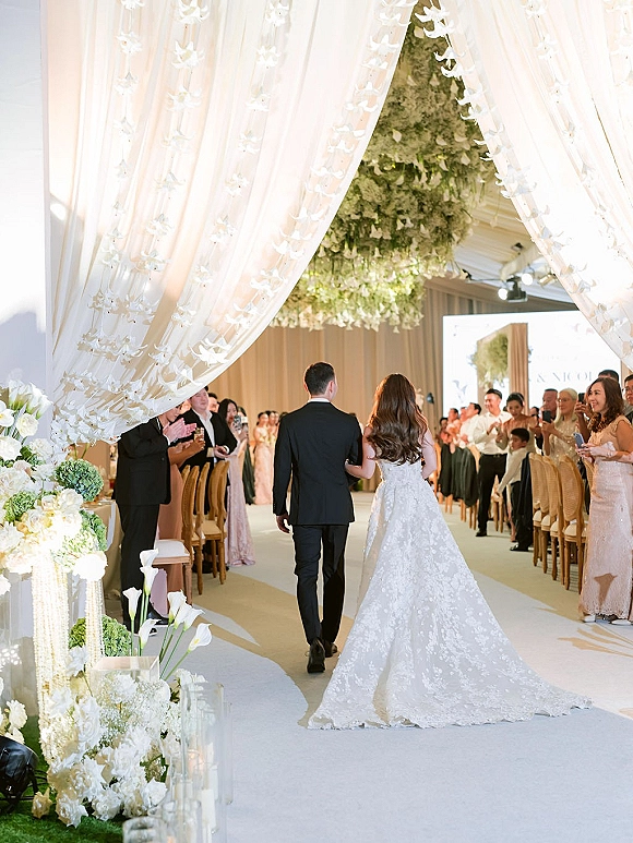 Wedding recessional as bride and groom walk down a candlelit aisle runner beneath hanging greenery, guests cheering at an indoor venue