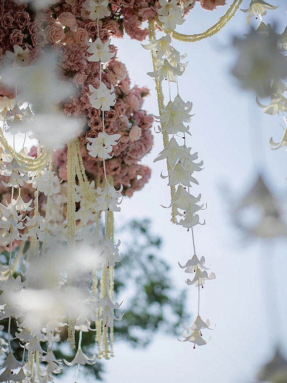 Hanging floral installation of pink roses and blush blooms with cascading white flower strands, suspended against blue sky and tree foliage