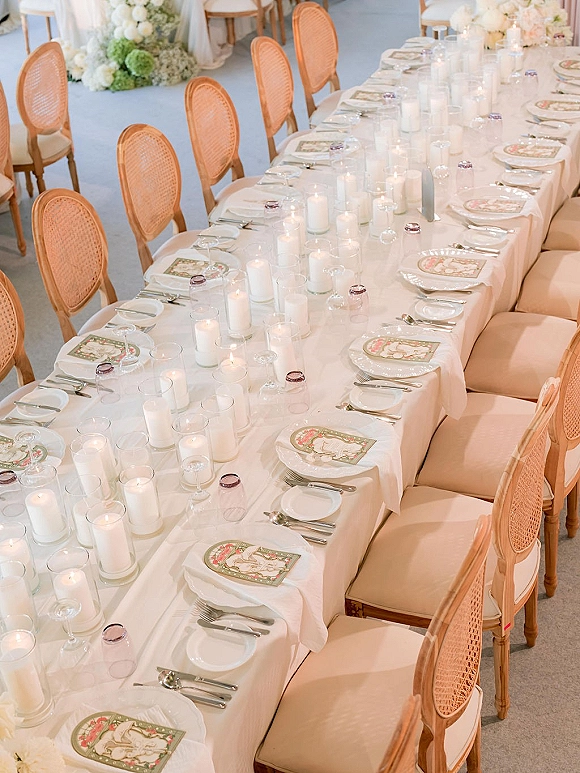 Reception tablescape on a long wedding table with white linen, taper and votive candles, glass vases, layered place settings, and florals before a draped backdrop