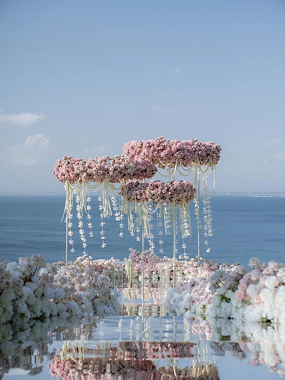 Wedding ceremony arch with pink and white hanging floral garlands over a reflective aisle runner, set against an ocean horizon backdrop