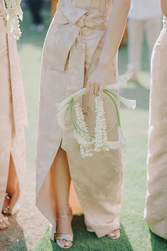 Bridesmaid bouquet of white calla lilies held against a satin dress bow, ring visible, on a sunlit lawn with greenery backdrop