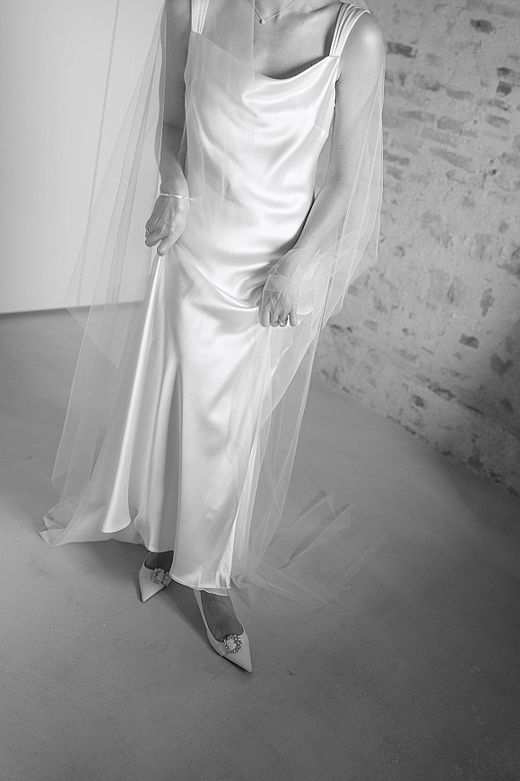 Wedding dress in satin slip style with sheer veil drape, paired with pointed toe heels and jewelry against a brick wall doorway backdrop
