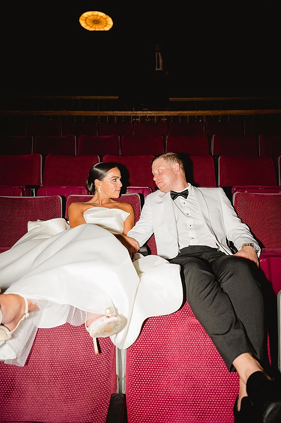 Couple portrait of bride and groom seated on theater seats, gazing at each other in a dark auditorium under a ceiling light fixture