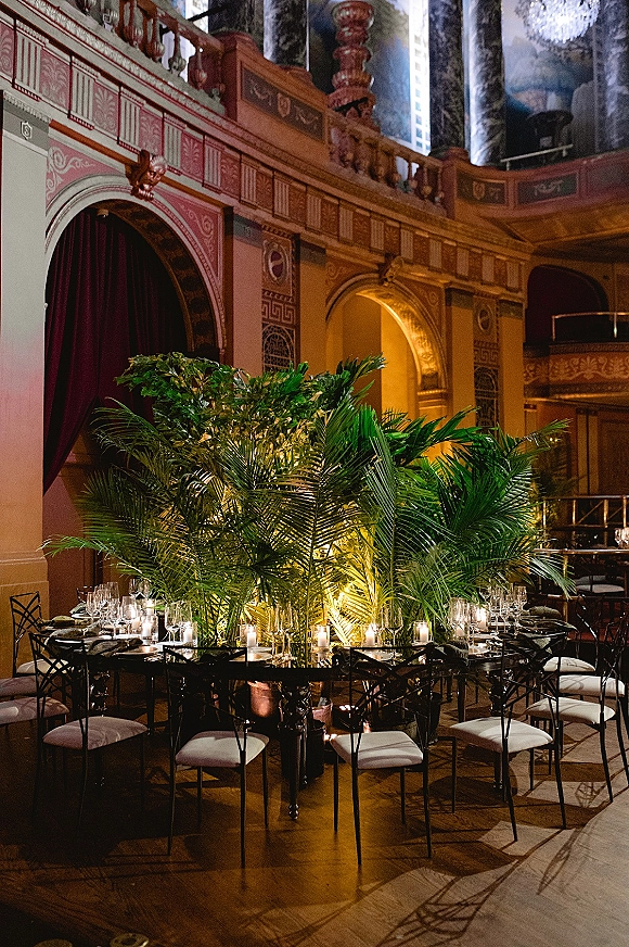 Reception tablescape with a palm leaf centerpiece, taper and votive candles, and glassware set in an ornate ballroom under a chandelier