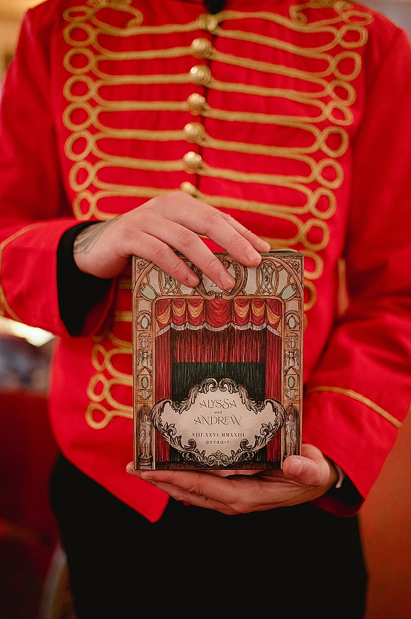 Invitation suite theatre wedding invitation displayed in a keepsake box beside a red military-style jacket with gold braid trim and buttons indoors