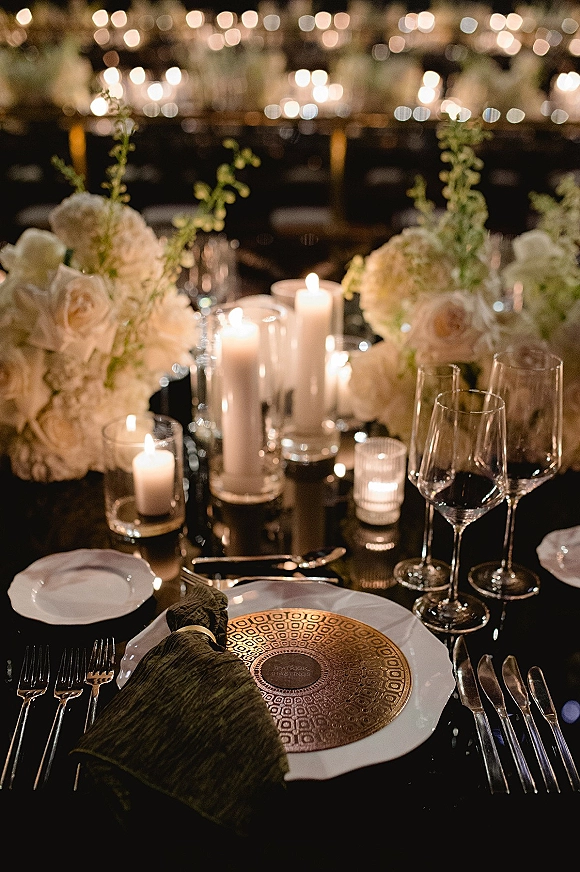 Reception tablescape with candlelit wedding table styling, white hydrangeas and roses, greenery, and gold chargers in a dim room with bistro lights