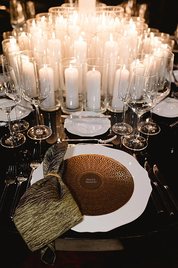 Reception tablescape with wedding candle centerpiece of white pillar candles in glass hurricanes, gold chargers and glassware on black linens under dim light