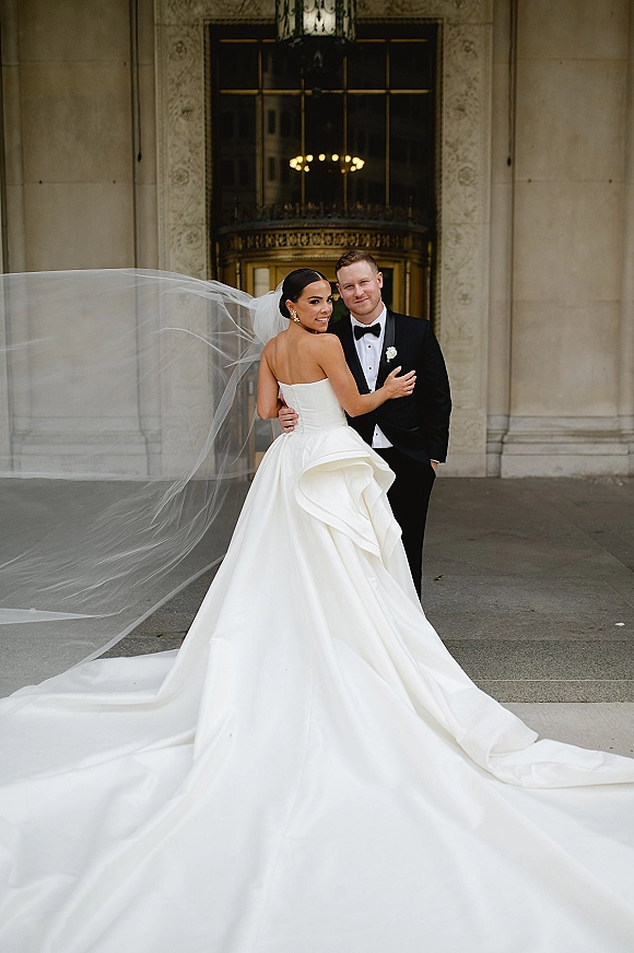 Couple portrait of bride and groom embrace at a stone building entrance, her long veil blowing behind a strapless gown train