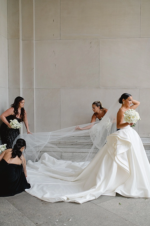 Bride portrait with long veil as bridesmaids in black dresses adjust her cathedral train on stone steps by a stone wall