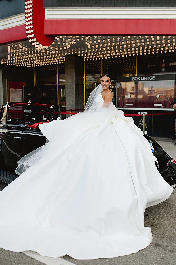 Bridal portrait of a bride in a strapless ball gown with cathedral veil, seated in a vintage convertible by theater marquee lights on a city street