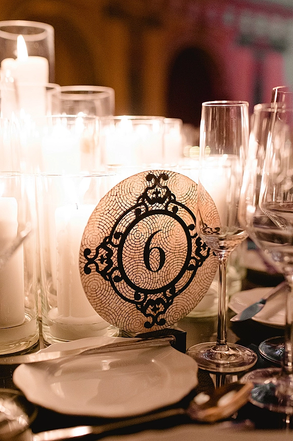 Wedding table number card in a black and ivory holder beside pillar candles in glass cylinders, champagne flute, and formal place setting under warm light