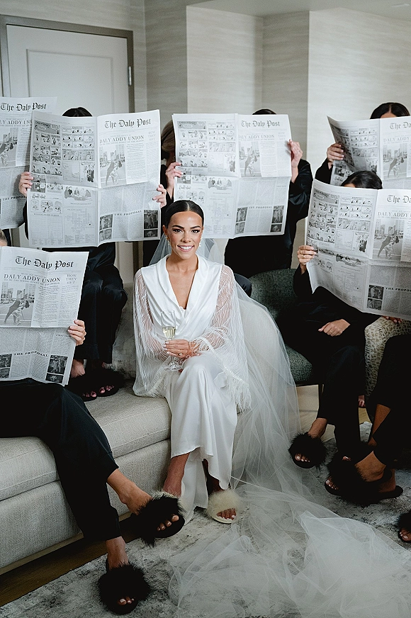 Bride portrait in a white robe with a long veil, holding a champagne flute on a sofa in a neutral hotel suite with bridesmaids in black pajamas holding newspapers