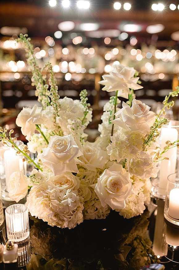 Wedding centerpiece with white rose centerpiece of hydrangeas and pillar candles on a mirrored table under bistro lights in the reception room