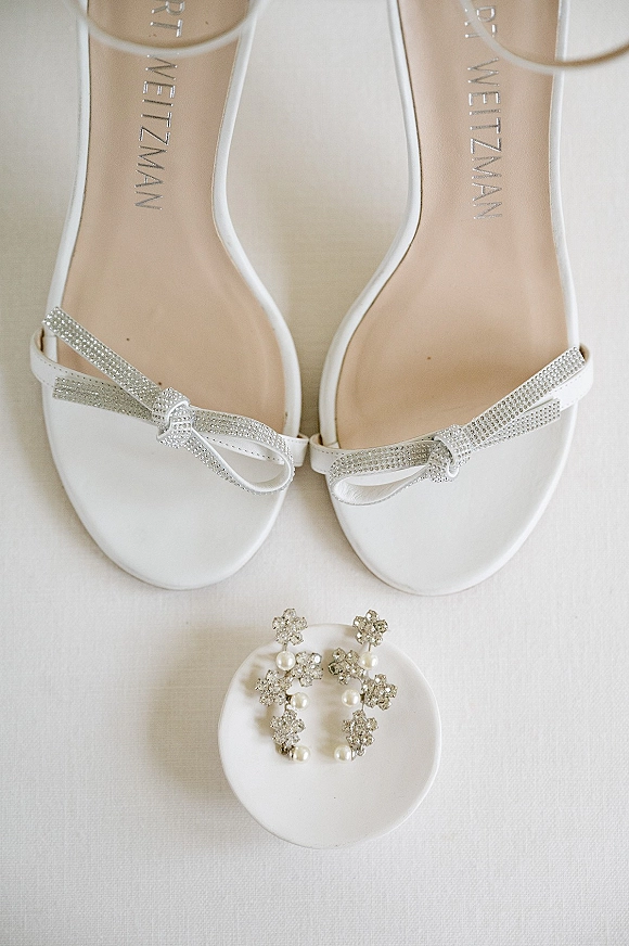 Bridal shoes flatlay with white wedding sandals featuring rhinestone bow straps, earrings and round ring box on white fabric backdrop
