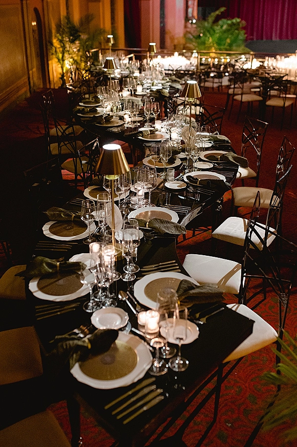 Reception tablescape with black wedding tablescape styling on a long banquet table, candlelit place settings, lamps, and greenery in a dim room