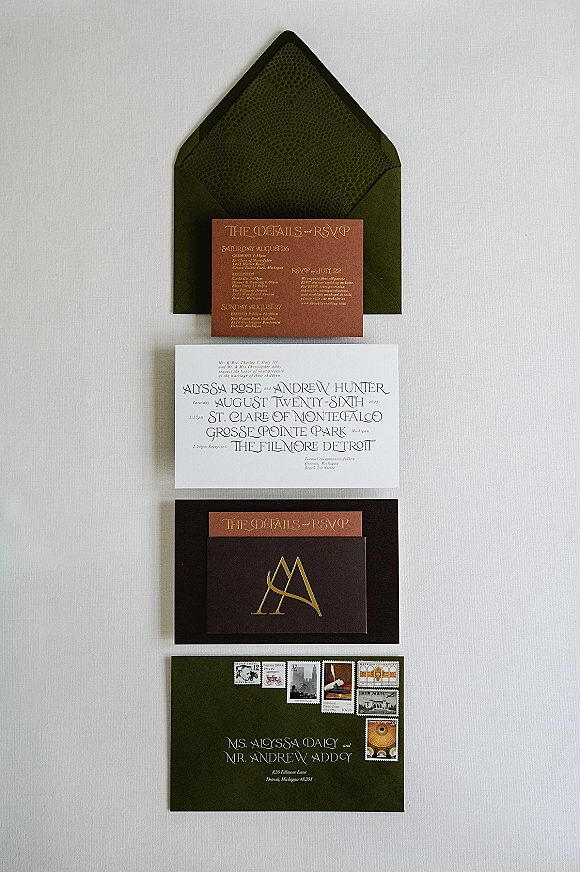 Wedding invitation suite with modern wedding invitations in dark green envelopes, gold foil lettering, stamps, and cards on light fabric backdrop