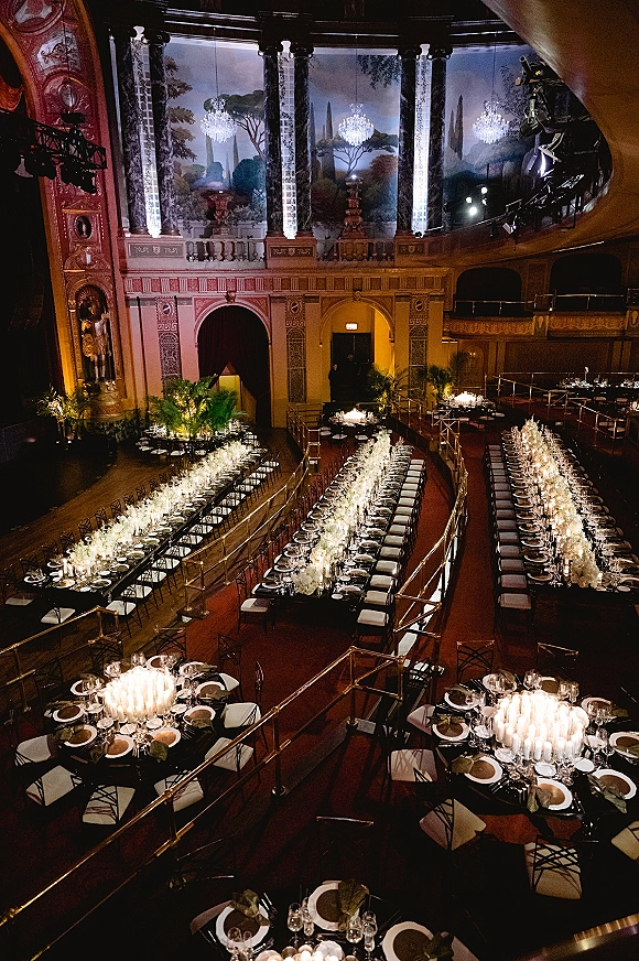 Reception tablescape with long banquet tables wedding styling, white floral centerpieces and candles under chandeliers in an ornate theater interior