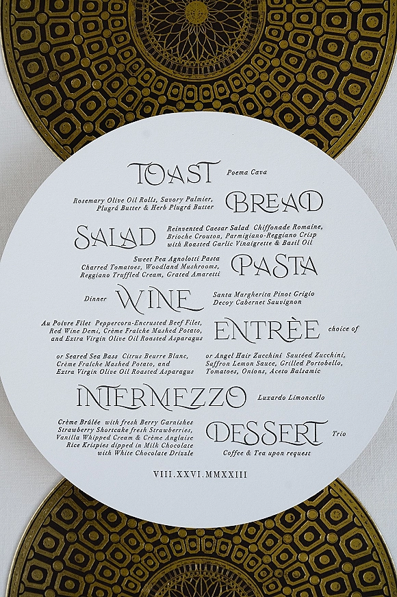 Wedding menu card on a gold patterned charger plate over neutral linen, featuring a round design with modern black-and-white typography