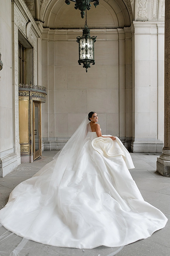 Bridal portrait of a bride in a strapless ball gown with cathedral veil and long train, looking back beneath a stone archway and lantern