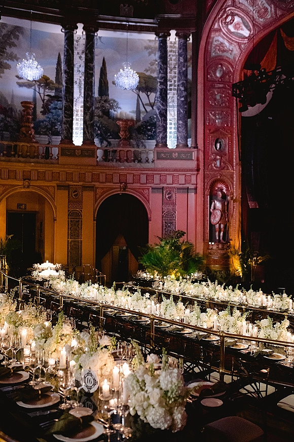 Reception tablescape on a long banquet table wedding with white floral centerpieces, taper candles, and chandeliers in an ornate theater interior