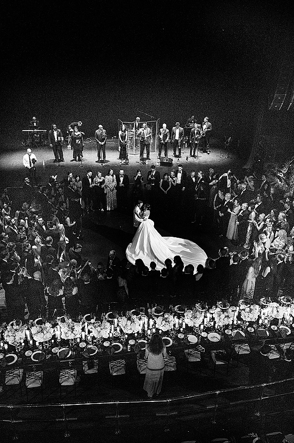 First dance with bride in long-train wedding gown and groom in tuxedo, guests encircling them, live band on stage in dark venue