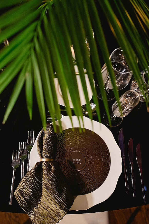 Reception place setting wedding tablescape with white charger, patterned plate, black napkin ring, wine glasses and palm leaves on dark table