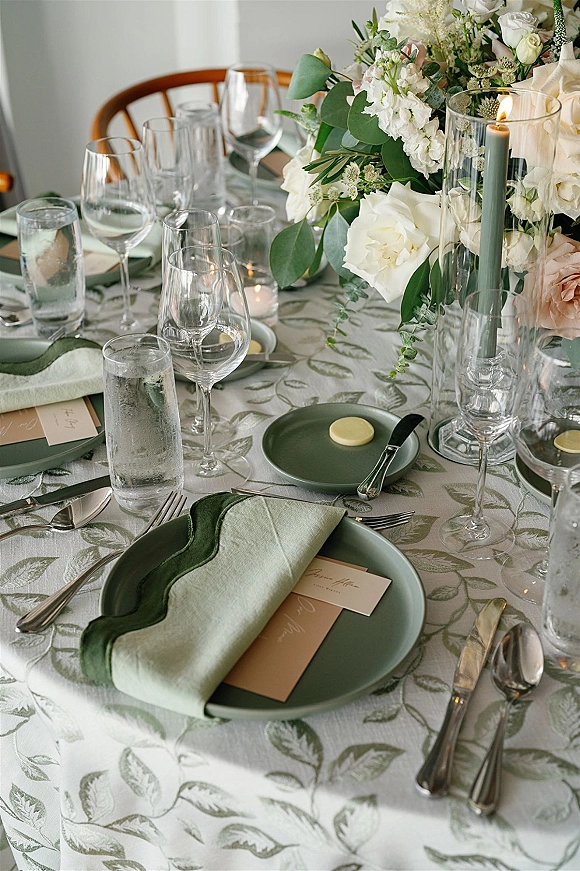 Reception tablescape wedding table setting with sage green plates, white rose centerpiece, taper candles in glass hurricanes on patterned cloth by a white wall