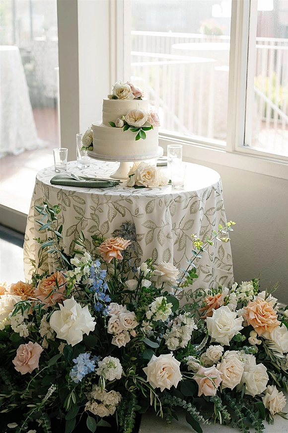 Wedding cake table with a two tier wedding cake, sugar roses and greenery on a stand, patterned cloth, votive candles by window light