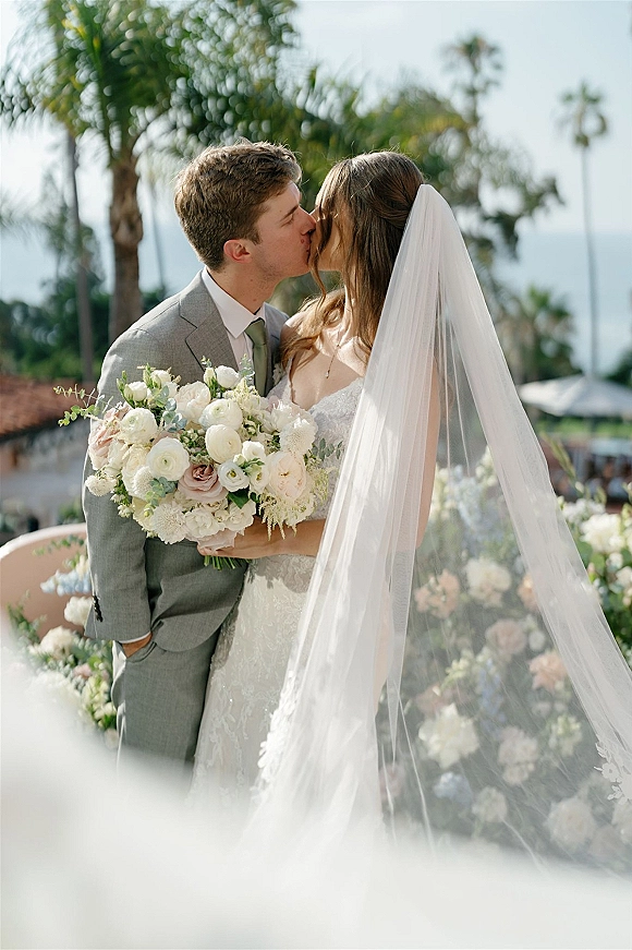 Wedding kiss portrait of bride and groom kissing, bride holding white and blush rose bouquet with eucalyptus on a palm-lined terrace by the ocean