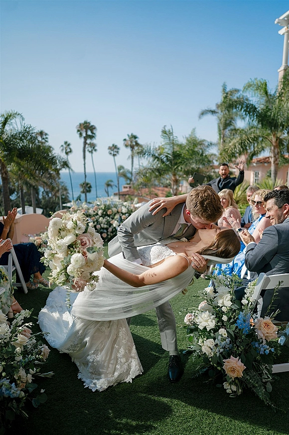 Wedding kiss moment as groom dips bride holding a bouquet, veil flowing down a floral aisle with guests, palms and ocean beyond