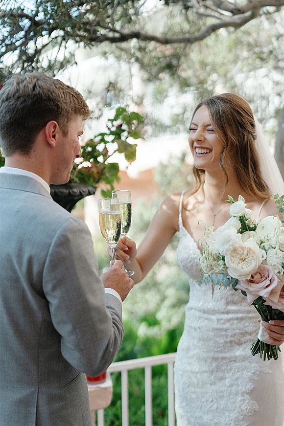 Wedding toast as bride and groom clink champagne flutes, bride holding rose bouquet in veil on an outdoor patio with trees in daylight