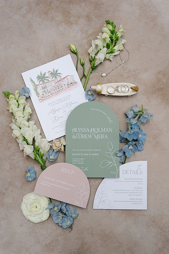 Invitation suite flat lay with save the date, RSVP and details cards beside engagement rings and flowers on a neutral stone surface