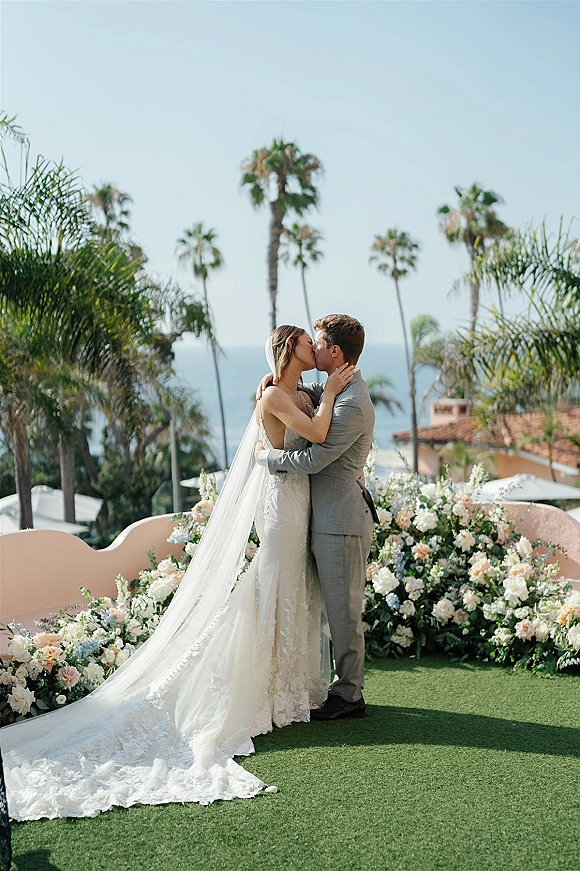 Wedding kiss as the bride and groom kissing on a coastal lawn, her long cathedral veil trailing by florals with palm trees and ocean view