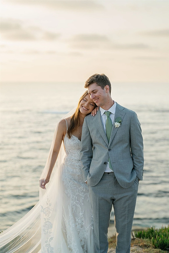 Couple portrait at the shoreline, bride leaning on groom in a strapless lace wedding dress and veil with ocean and clouds behind them