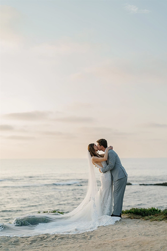 Wedding kiss portrait of bride and groom kissing on the beach shoreline, with a long veil flowing over sand and coastal rocks behind