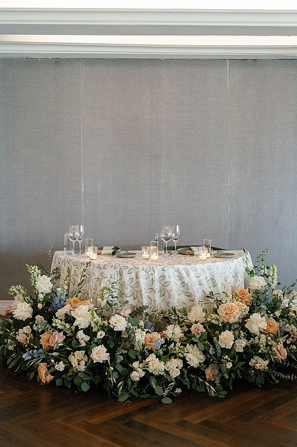 Sweetheart table decor with sweetheart table flowers, roses and greenery on the floor, votive candles, and place settings against gray wall panels