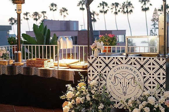 Wedding churro bar wedding dessert station with patterned tile cart, heat lamp, wicker baskets and florals on a sunset rooftop terrace with ocean view
