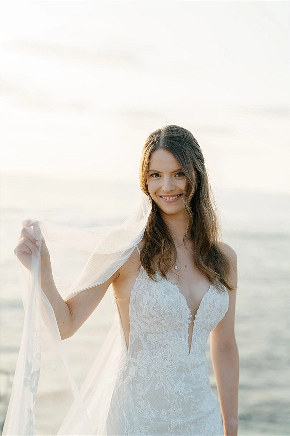 Bridal portrait of a bride holding veil, wearing a lace deep V wedding dress and necklace, smiling with ocean horizon behind her