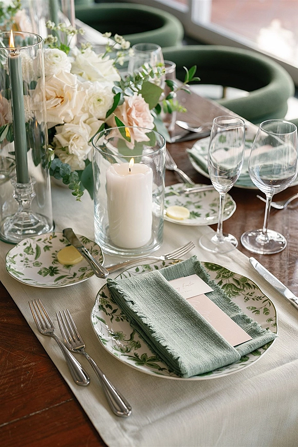 Reception tablescape with green and white tablescape details, blush and white roses, sage napkins, candlelight, and place settings on a wood table by window light