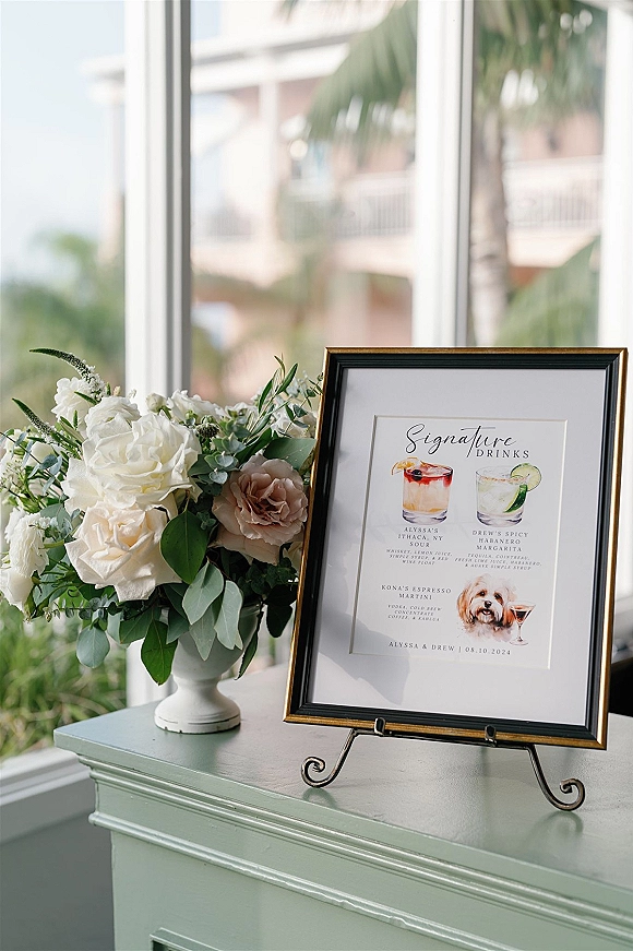 Signature drinks sign with wedding signature cocktails in a black and gold frame on an easel, beside rose and greenery florals by a sunlit window