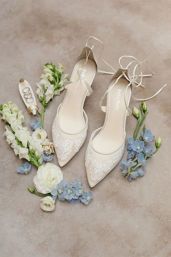 Wedding shoes flatlay featuring lace wedding shoes with ankle-wrap ribbons, rings and velvet ring box, plus blue and white flowers on stone background