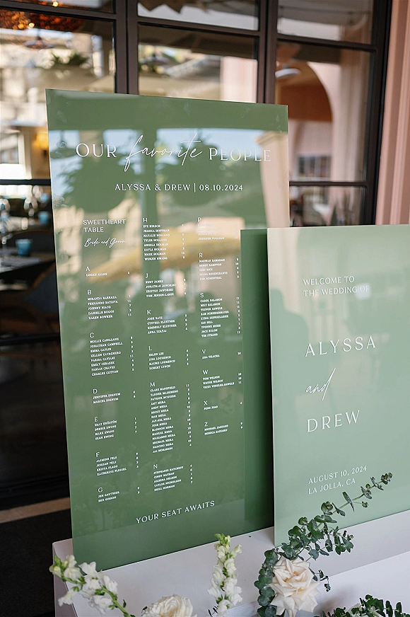 Wedding seating chart on an acrylic seating chart sign with white lettering, green panels, and white roses with greenery by glass windows indoors