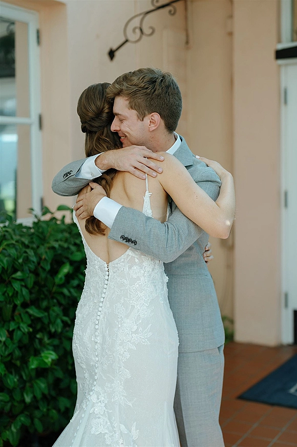 First look moment as bride in a backless lace wedding dress hugs groom in a gray suit by a stucco doorway with greenery hedge