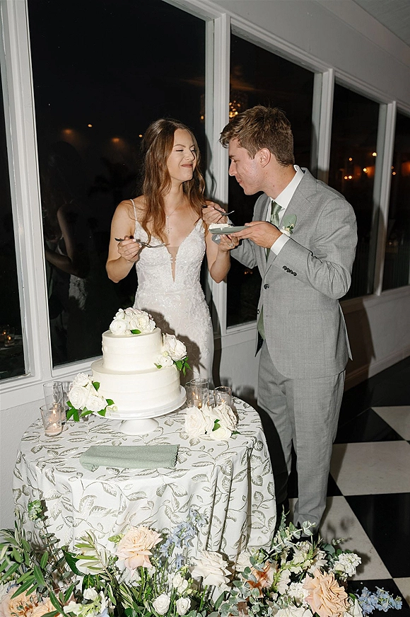 Wedding cake cutting as bride in lace dress and groom in suit slice a two-tier white cake with floral topper and candlelit table by tall windows at night