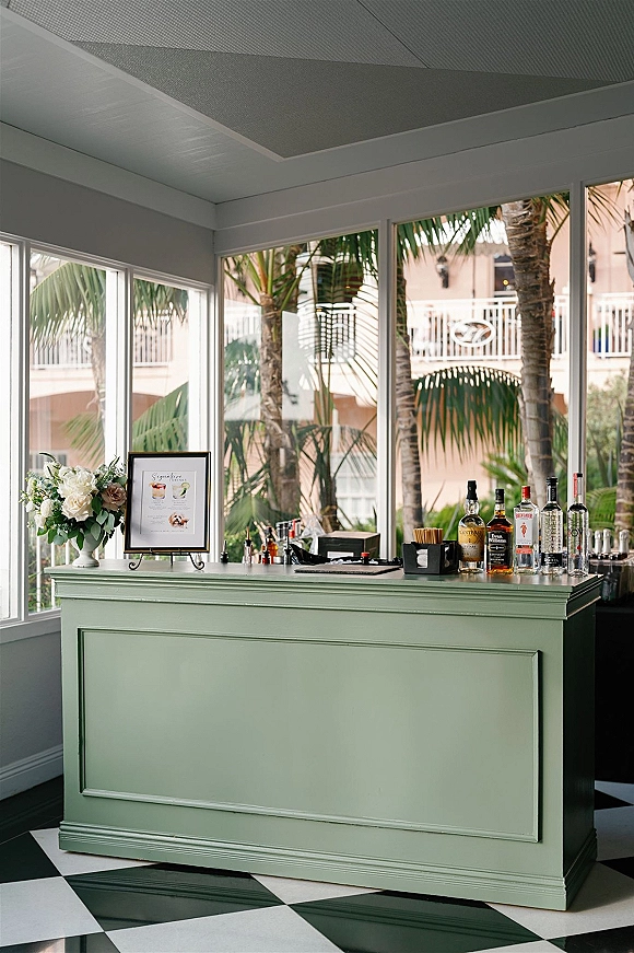 Wedding bar setup with mobile wedding bar counter displaying liquor bottles, tools, and a floral arrangement on a black-and-white floor by tall windows