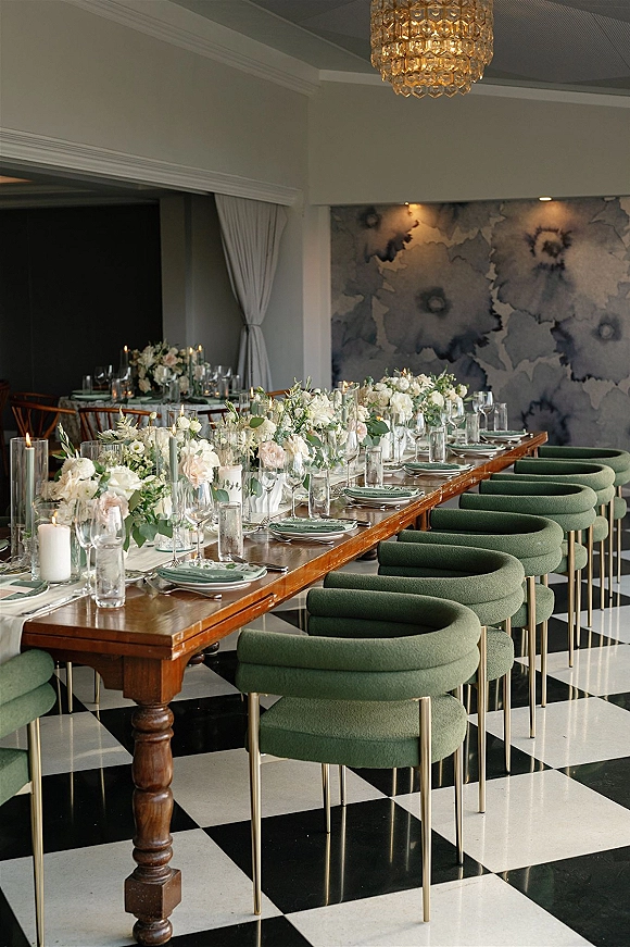 Reception tablescape with a long banquet table, white floral centerpieces and pillar candles, set on a black-and-white checkered floor under a chandelier