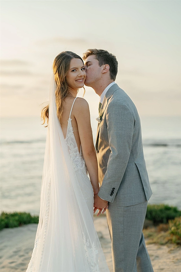 Wedding couple portrait of groom kissing the bride’s cheek as they hold hands, her long veil and lace dress flowing by the ocean coastline