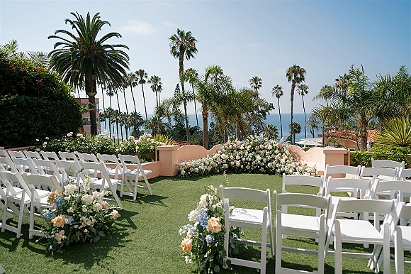 Ceremony setup with outdoor wedding ceremony seating of white folding chairs and floral aisle markers on a coastal lawn with ocean view and palms