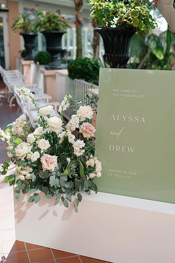 Wedding welcome sign on a pedestal with a white and blush rose floral arrangement and eucalyptus on an outdoor patio entrance setup