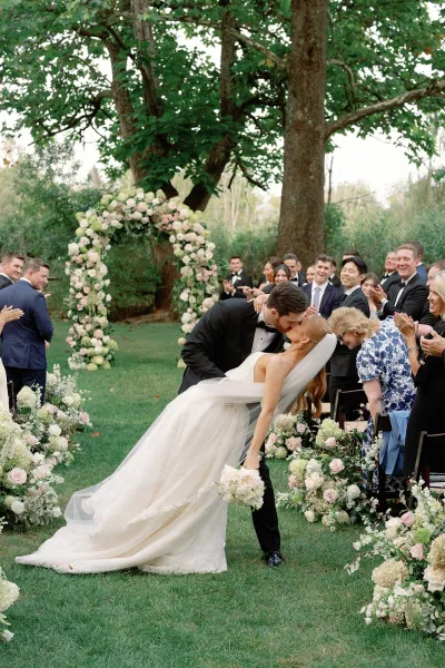 Wedding kiss portrait of bride and groom in a dip, veil flowing as she holds a bouquet beneath a floral arch on a green lawn under trees